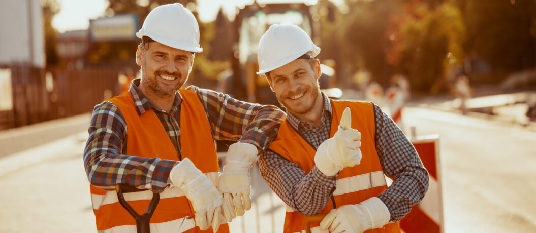 Two construction workers in hardhats and vests posing for a photo at the road